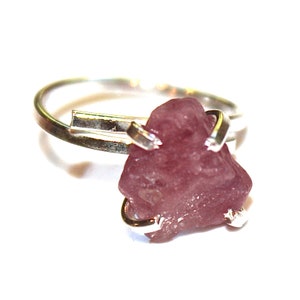 May include: A silver ring featuring a pink, rough-cut gemstone set in the centre. The ring is made of two bands that cross over each other.