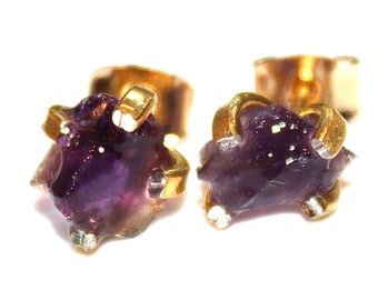 May include: A pair of gold stud earrings with purple amethyst gemstones. The amethyst is raw and has a natural, irregular shape.
