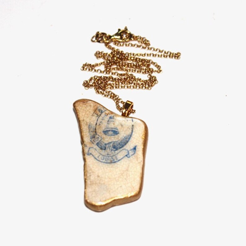 May include: A gold chain necklace with a pendant made from a piece of ceramic tile. The tile has a blue and white design with the words "The" and "Tourist" on it.