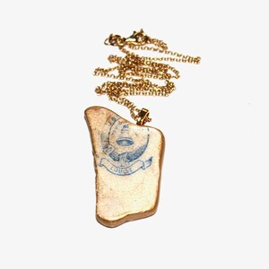 May include: A gold chain necklace with a pendant made from a piece of ceramic with a blue and white design. The design features a stylized eye with a banner that reads "L'OISE."
