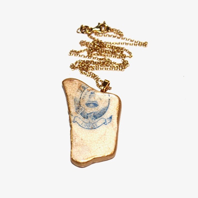 May include: A gold chain necklace with a pendant made from a piece of ceramic with a blue and white design. The design features a stylized eye with a banner that reads "L'OISE."