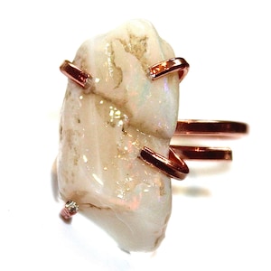 May include: A ring featuring a large, white opal stone set in a copper wire frame. The opal has a natural, rough texture and a subtle iridescent sheen. The ring is a unique and eye-catching piece of jewellery.