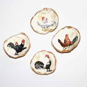 May include: Four decorative oyster shell dishes with gold trim, each featuring a different rooster design. The roosters are depicted in various colors, including white, black, and red.