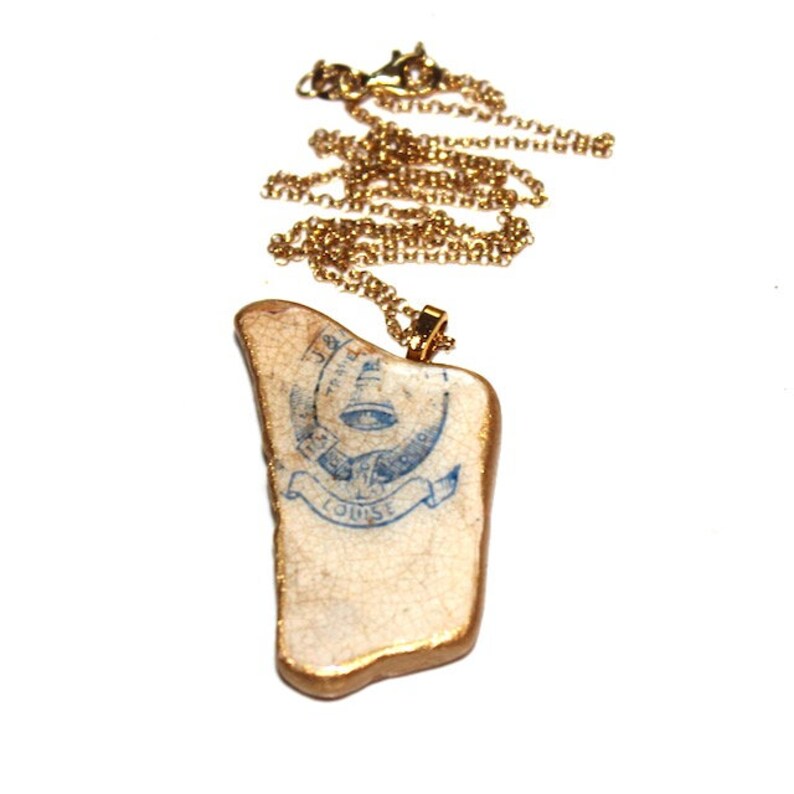 May include: A gold chain necklace with a pendant made from a piece of ceramic with a blue and white design. The design features a bell, a banner with the word "LOUISE" and the text "The Midland Bell Co."