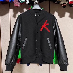 Könnte beinhalten: Eine schwarze Collegejacke mit schwarzen Lederärmeln, einem schwarzen Wollmischgewebe-Körper und einem auffälligen roten Buchstaben K als Aufnäher auf der linken Brust. Die Jacke hat eine Druckknopfleiste und zwei Seitentaschen mit Lederbesatz.