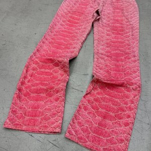 May include: Bright pink pants featuring a textured, embossed snakeskin pattern throughout the fabric. These trousers have a standard waistband with a button closure, a front zipper fly, and a straight-leg cut that extends down to the hemline.