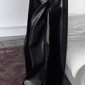 May include: Black wide-leg trousers featuring a glossy, textured crocodile-skin pattern. The trousers have a high-waisted fit and a long, straight-leg silhouette that extends down to the floor, with a subtle sheen across the dark material.