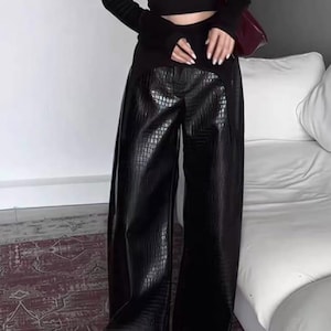 Puede incluir: Un top corto negro de manga larga combinado con pantalones de tiro alto y pernera ancha con una textura negra brillante de efecto cocodrilo. El conjunto se completa con un bolso de hombro rojo oscuro con hebilla plateada y un collar de corazón.