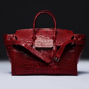 May include: A deep red handbag featuring a textured crocodile skin pattern. The bag includes two top handles, a central flap closure, and a decorative horizontal strap across the front secured with a small silver-toned metal buckle.