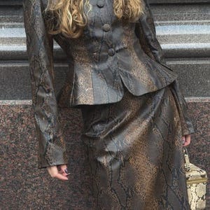 May include: A two-piece set consisting of a long-sleeved jacket and a matching skirt, both featuring a brown and black snakeskin pattern. The jacket has a high collar, a button-front closure, and a peplum hem. A small, structured box bag is included.
