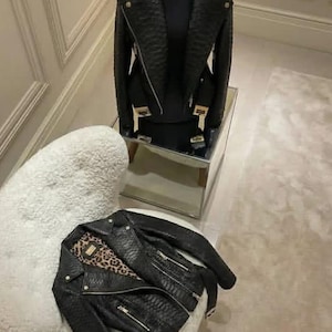 May include: A black textured leather motorcycle jacket with silver-tone hardware, featuring a wide notched collar, asymmetrical front zipper, and a leopard print lining. The jacket is displayed on a mannequin and also laid flat.
