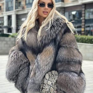 Könnte beinhalten: Ein dicker, langärmeliger Kunstpelzmantel mit einem Farbverlauf in Grau-, Silber- und Brauntönen und einem großen Kragen. Der Mantel wird mit einer strukturierten Clutch mit grau-weißem Schlangenledermuster und dunkel getönten Sonnenbrillen kombiniert.