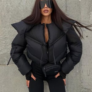 Peut inclure: Une doudoune courte noire avec un col montant, des manches longues et une fermeture éclair frontale. La veste présente un sac banane noir assorti attaché à la taille. La tenue est complétée par un pantalon noir ajusté et des lunettes de soleil visière.