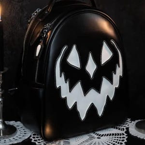 Könnte beinhalten: Ein schwarzer Rucksack mit einem großen, weißen, gezackten Kürbisgesicht auf der Vorderseite. Die Tasche hat eine abgerundete Oberseite, einen Tragegriff und silberfarbene Reißverschlüsse. Das Design zeigt zwei dreieckige Augen und ein breites Grinsen.