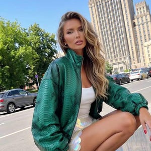 May include: A vibrant emerald green jacket featuring a textured snakeskin pattern, a high collar, and a full-length front zipper. The jacket has long sleeves with elasticized cuffs and is worn over a simple white top and a white iridescent skirt.