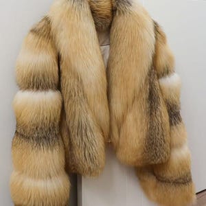 May include: A long-sleeved jacket made of thick, multi-toned fur in shades of golden tan, cream, and dark brown. The garment features a wide, plush collar and horizontal bands of fur along the sleeves, creating a voluminous and textured appearance.