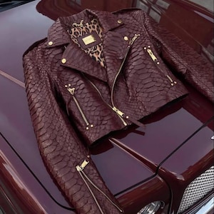 May include: A dark burgundy motorcycle jacket with a textured snakeskin-like pattern. The jacket features gold-toned metal zippers on the front, sleeves, and pockets, along with matching gold buttons. The interior lining displays a brown leopard print.