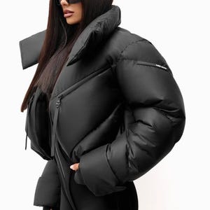 Könnte beinhalten: Eine schwarze Daunenjacke mit hohem Kragen, langen Ärmeln und einem diagonalen Reißverschluss auf der Vorderseite. Die Jacke hat eine matte Oberfläche, breite Steppnähte und ein kleines rechteckiges Etikett am linken Ärmel mit dem Text MISS WOW.