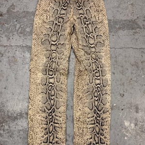 May include: Full-length trousers featuring a prominent snakeskin pattern in shades of beige, tan, and dark brown. The design includes a vertical, darker-toned scale motif running down the center of each leg, creating a symmetrical appearance.