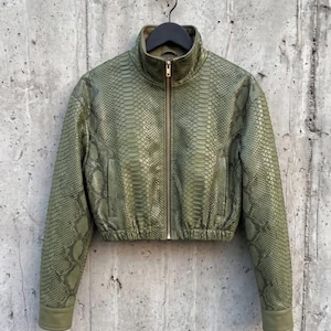 May include: A cropped jacket in a muted olive green color featuring a textured snakeskin pattern throughout. The garment has a high collar, a central gold-toned zipper closure, long sleeves with solid cuffs, and an elasticized hem at the waist.