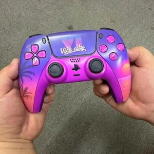 Può includere: Un controller per videogiochi wireless con una sfumatura di colore viola e rosa. Il touchpad centrale mostra il testo Vice City in un carattere stilizzato. Il controller include levette analogiche nere, pulsanti azione rosa e impugnature decorate con sagome di palme e un tramonto.
