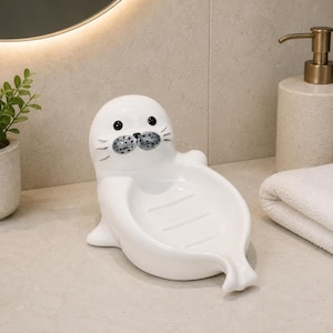 Peut inclure: Un porte-savon en céramique blanche en forme de phoque. Le phoque présente une finition lisse et brillante avec des yeux peints en noir et un museau gris tacheté. Le plat comporte un centre concave avec trois rainures pour maintenir le savon.