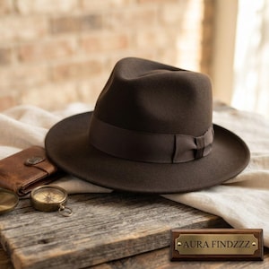 Könnte beinhalten: Ein dunkelbrauner Fedora-Hut mit einem passenden braunen Ripsband. Der Hut hat eine eingedrückte Krone und eine breite Krempe. Eine kleine rechteckige Holzplakette mit dem Text AURA FINDZZZ befindet sich in der unteren rechten Bildecke.