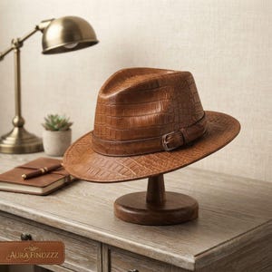 Peut inclure: Un chapeau fedora marron clair avec un motif texturé en peau de crocodile, une calotte structurée et une bande en cuir assortie dotée d'une boucle en métal argenté. Le chapeau est présenté sur un support en bois, révélant sa surface gaufrée.