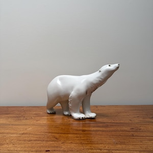 Puede incluir: Una figura de oso polar de porcelana blanca de pie en una pose orientada hacia adelante con la cabeza inclinada hacia arriba. El oso presenta un acabado liso y brillante, patas moldeadas detalladas y contornos anatómicos sutiles en su postura.