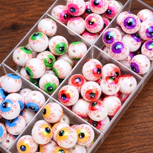 Op de afbeelding: Een verzameling bolvormige kralen die lijken op oogbollen, elk met een witte basis met rode aders en een centrale iris in verschillende kleuren, waaronder roze, paars, groen, blauw en geel, georganiseerd in een doorzichtige plastic bak.