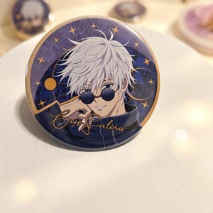 Op de afbeelding: Een ronde button met een anime-personage met wit haar en een ronde zwarte zonnebril. De achtergrond is een donkerblauwe nachtelijke hemel met sterren en een gouden cirkel. De tekst Gojo Satoru is geschreven in een cursief lettertype.