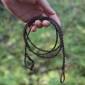 May include: A dark brown, multi-strand braided cord necklace or accessory featuring a twisted rope-like texture. The cord is formed into a circular shape with two small loops at the ends, showcasing a consistent dark brown colour throughout the length.