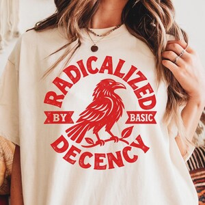 Pode incluir: Uma t-shirt de manga curta na cor creme com uma estampa gráfica vermelha de um corvo pousado num galho. O texto RADICALIZED BY BASIC DECENCY está impresso numa fonte vermelha circular em torno da ilustração do pássaro.