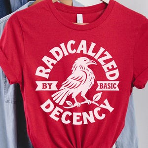 Könnte beinhalten: Ein rotes Kurzarm-T-Shirt mit einer weißen Grafik eines auf einem Ast sitzenden Raben. Der Text "RADICALIZED BY BASIC DECENCY" ist kreisförmig um den Vogel gedruckt. Das T-Shirt ist am unteren vorderen Saum zu einem Knoten gebunden.