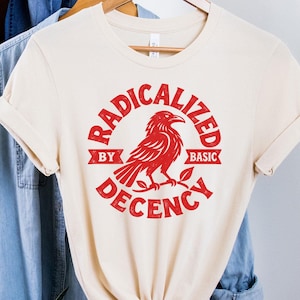 Puede incluir: Una camiseta de manga corta color crema con un gráfico rojo de un cuervo posado en una rama. El texto RADICALIZED BY BASIC DECENCY está impreso en una fuente roja circular alrededor del ave. La camiseta está anudada en el dobladillo.