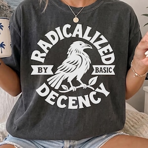 Może przedstawiać: Ciemnoszary t-shirt z krótkim rękawem z białą grafiką przedstawiającą kruka siedzącego na gałęzi. Napis RADICALIZED BY BASIC DECENCY jest nadrukowany w okrągłym wzorze wokół ptaka. Koszulka ma okrągły dekolt i luźny krój.