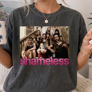 Puede incluir: Una camiseta gris carbón que presenta un gráfico rectangular del elenco de la serie de televisión Shameless, con la palabra Shameless impresa en rosa debajo de la imagen. La camiseta tiene cuello redondo y mangas cortas, combinada con shorts.