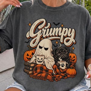 Könnte beinhalten: Ein anthrazitfarbenes T-Shirt mit einem Grafikdruck, der den Text "Grumpy" in einer stilisierten Schrift zeigt. Das Motiv zeigt ein Gespenst und einen schwarzen Mops, die zusammen unter einer orange-schwarzen karierten Decke sitzen und Tassen halten.