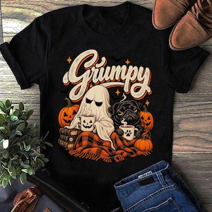 Puede incluir: Una camiseta negra con un estampado gráfico de un fantasma y un pug negro sentados juntos bajo una manta de cuadros naranja y negra. Ambos sostienen tazas con caras de calabaza. La palabra Grumpy está impresa en letras blancas grandes.