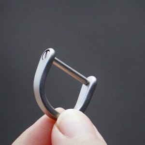 Pode incluir: Um anel de metal em forma de D na cor prata fosca com uma barra horizontal reta no centro. A estrutura curva tem um acabamento metálico escovado suave com um pequeno detalhe de parafuso circular visível na parte superior.