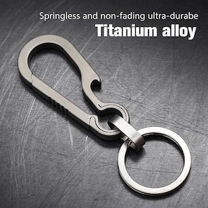 May include: A grey titanium alloy carabiner keychain featuring a springless design and an integrated bottle opener. The metal clip has a matte finish with textured grip detailing on the side and is attached to a circular split ring for keys.