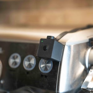 May include: A small black square-shaped electronic device with a single circular button and a tiny illuminated green light, mounted onto the metallic control panel of a stainless steel espresso machine featuring three circular silver buttons.
