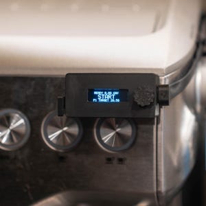 May include: A black, rectangular 3D-printed digital controller module attached to a stainless steel espresso machine. The device features a small blue backlit screen displaying the text READY 0.0G 60S START P1 TARGET 28.5G and a textured dial.