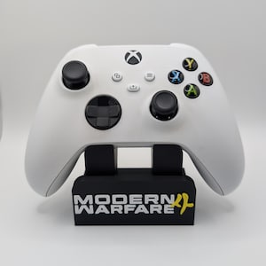May include: A white wireless video game controller with black joysticks and coloured action buttons, resting on a black plastic display stand featuring the white text "MODERN WARFARE" and a yellow logo on the front base of the stand.