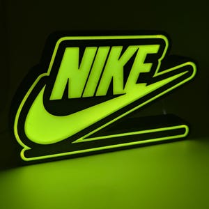 May include: A glowing neon-style sign featuring the Nike logo and text in a vibrant lime green colour. The sign has a black border and emits a bright light, displaying the word NIKE above the iconic swoosh symbol against a dark background.