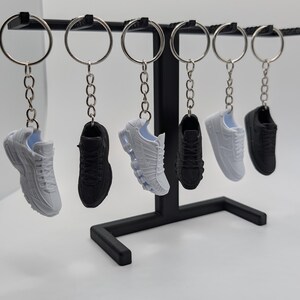 May include: Six miniature sneaker keychains hanging from a black metal display stand. The collection includes three white and three black shoe models, each attached to a silver-toned split ring by a short metal chain, showcasing detailed textures.