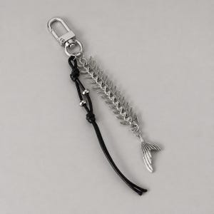 May include: A silver-toned metal keychain featuring a lobster clasp, a fish skeleton charm with a detailed tail, and a black leather cord accented with two small silver-toned metal beads, all attached to a central circular metal ring.