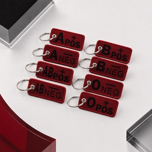 May include: A collection of eight rectangular red fabric keychains, each featuring a silver metal ring and black embroidered text representing blood types: A POS, A NEG, B POS, B NEG, AB POS, AB NEG, O POS, and O NEG, arranged in two columns.