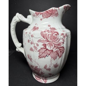 Pode incluir: Uma jarra de cerâmica branca com um padrão floral vermelho detalhado em transferência. O recipiente possui uma alça curva com detalhes moldados ornamentados, um bico largo e um corpo canelado. A estampa floral cobre a superfície externa.
