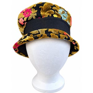 May include: A bucket hat featuring a black base with a raised, textured floral pattern in shades of mustard yellow, bright pink, and light blue. The hat includes a wide, solid black fabric band encircling the crown above the brim.